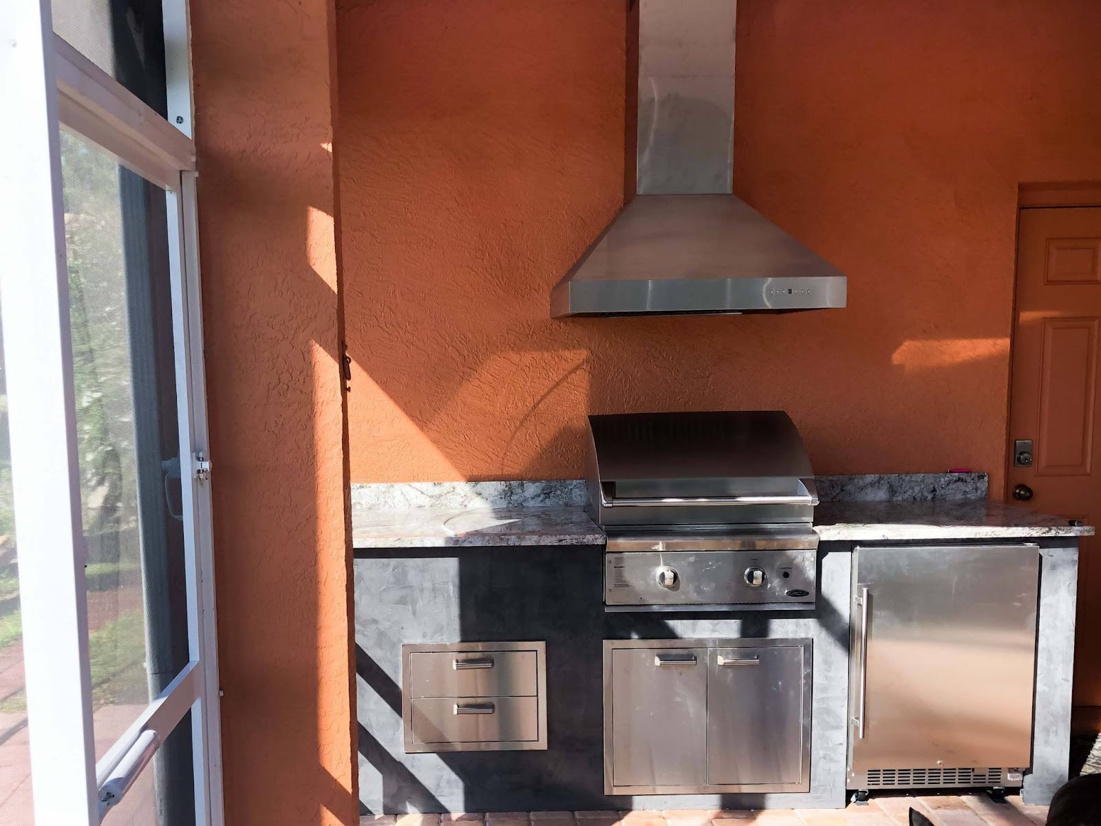 Compact outdoor kitchen with a stainless steel Proline range hood, grill, and storage on a screened-in porch with orange walls - prolinerangehoods.com