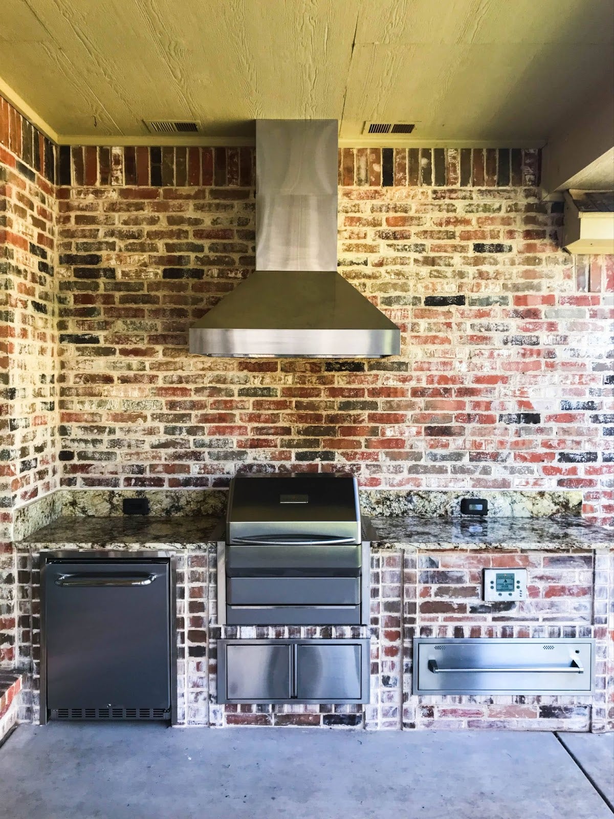 Outdoor kitchen with a Proline range hood, stainless steel appliances, and a rustic red brick wall - prolinerangehoods.com