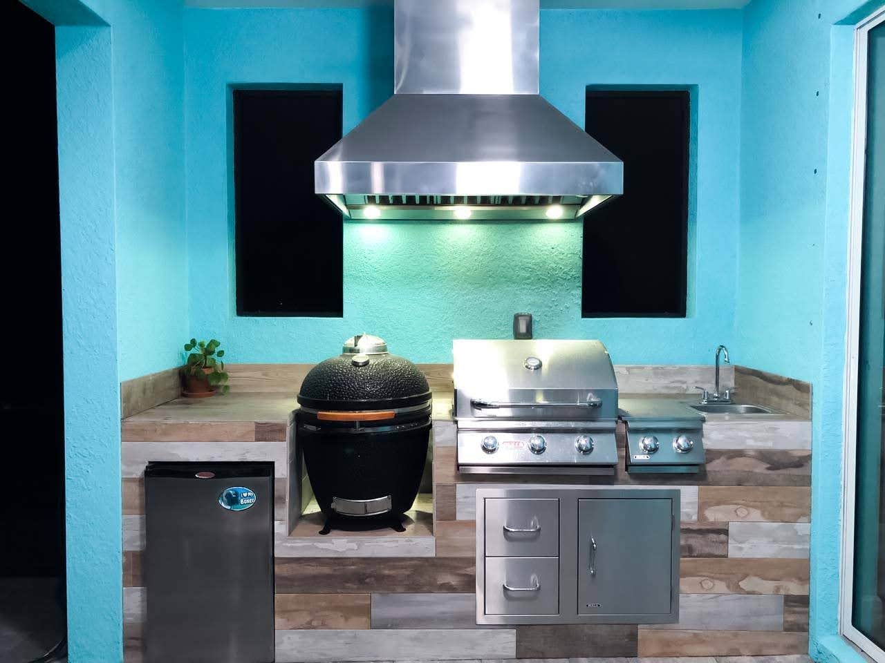 Contemporary outdoor kitchen featuring a Proline range hood, stainless steel grill, and ceramic smoker against a vibrant blue backdrop - prolinerangehoods.com