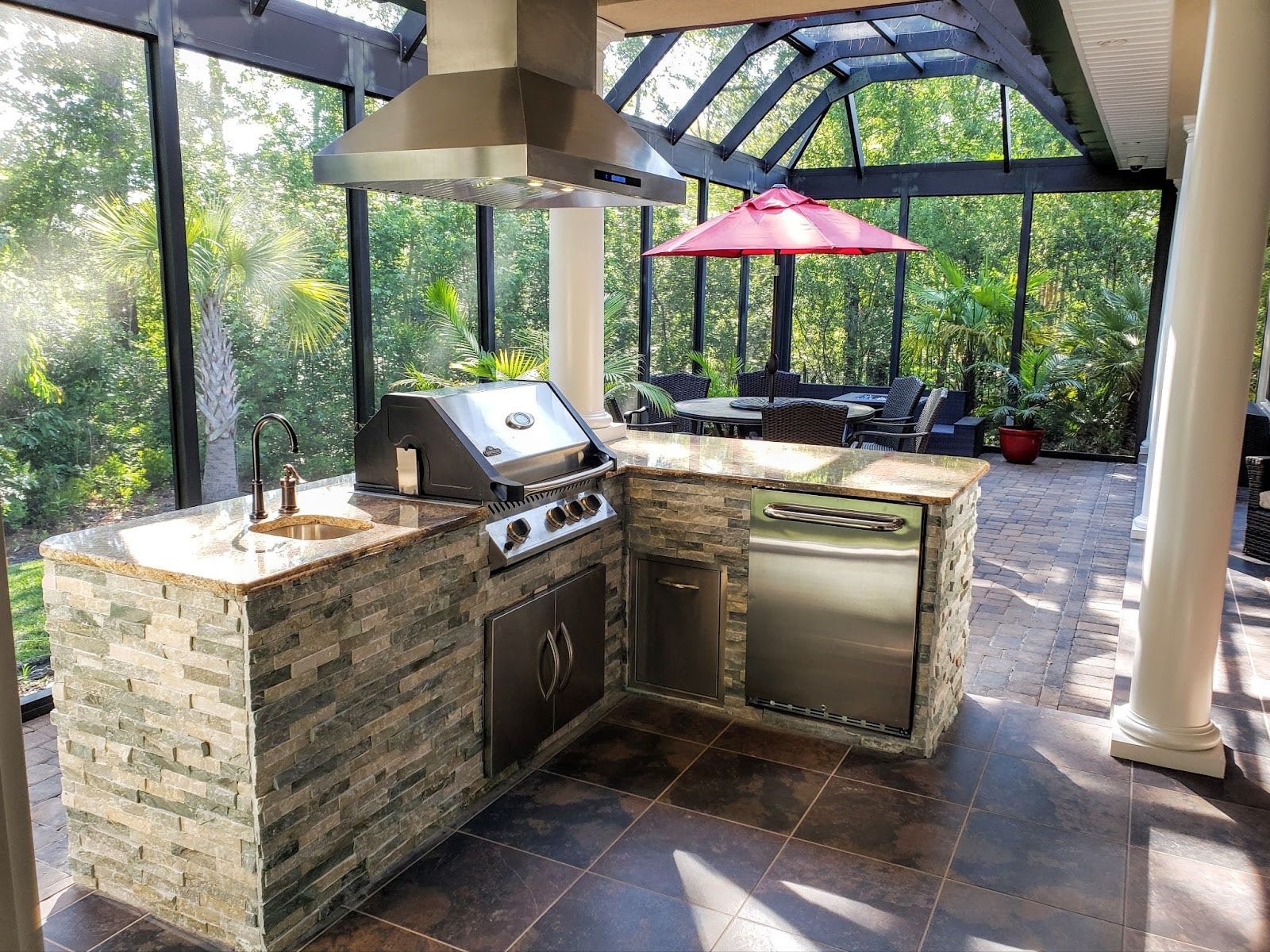 Luxurious outdoor kitchen with stainless steel appliances and stone cladding, complemented by a red umbrella patio set in a screened enclosure with a lush greenery backdrop.
