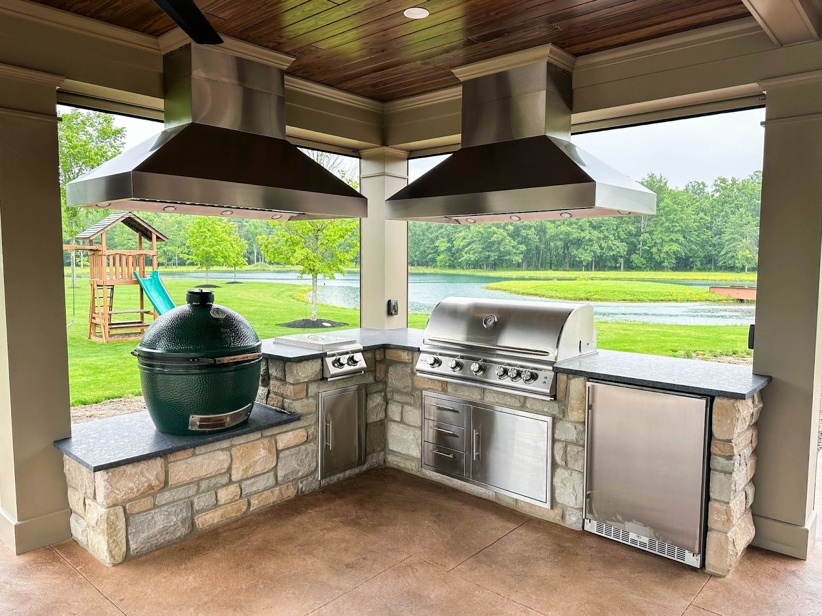 Spacious outdoor kitchen with premium stainless steel grill, smoker, and appliances set against a stone countertop with a view of a playground and pond in the background.