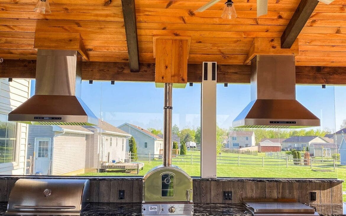 Stylish outdoor culinary setup with wood-paneled ceiling, featuring premium grilling equipment, ventilation hoods, and a view of a residential neighborhood.