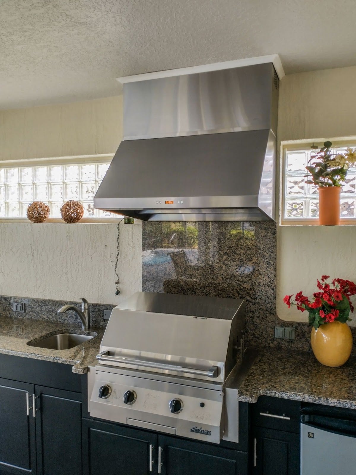 Cozy fun outdoor kitchen featuring a Proline wall range hood above a stove, framed by granite counters and decorative plants, with natural light from a glass block window - prolinerangehoods.com