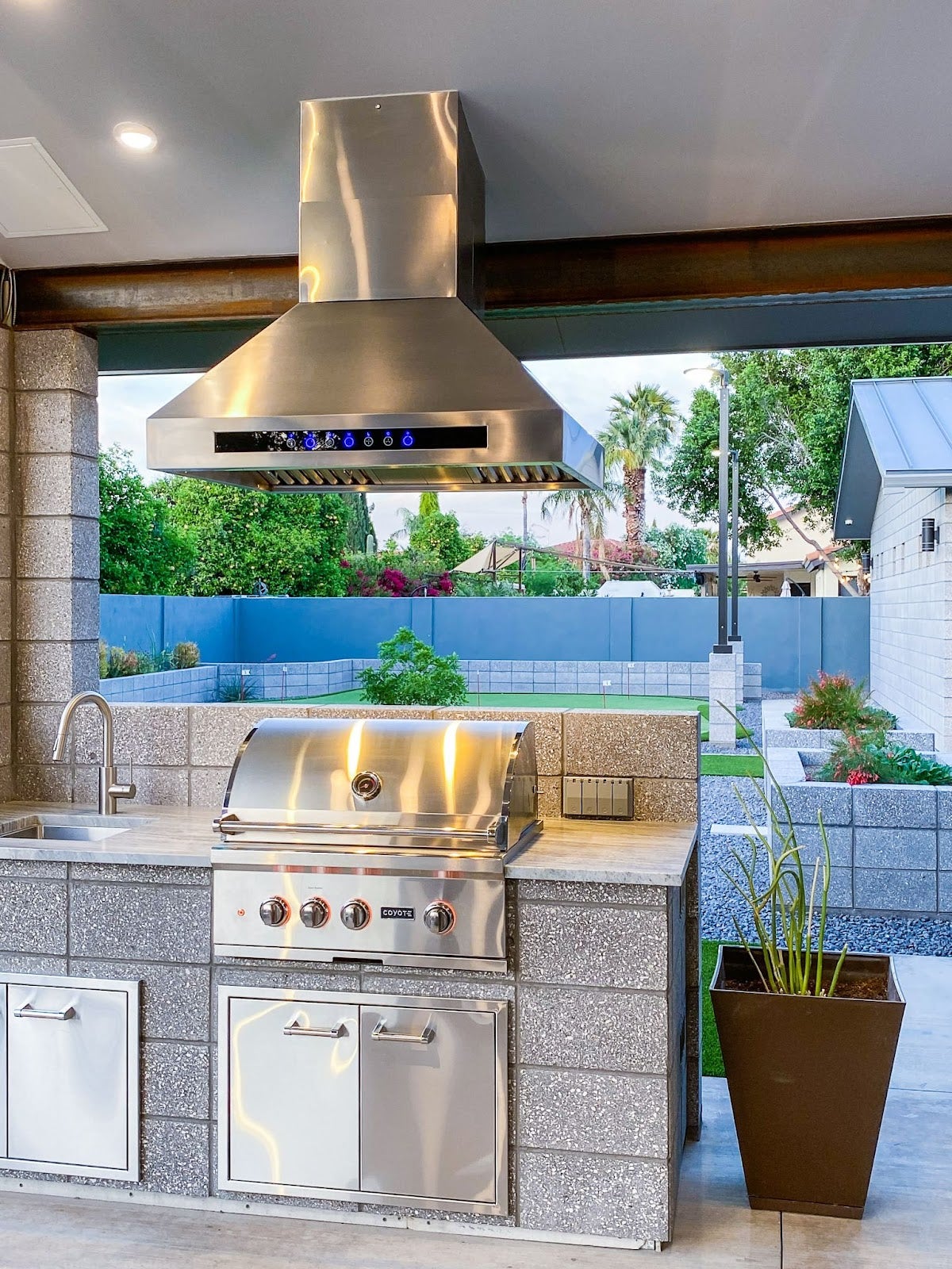 Elegant outdoor BBQ area featuring a high-end grill and stainless hood, nestled in a suburban oasis with lush greenery and modern landscaping.