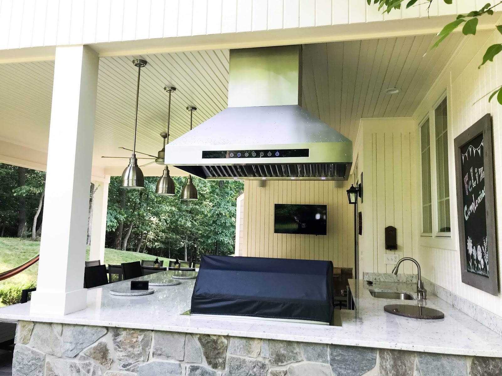 Charming outdoor entertaining area complete with a modern grill under a ventilation hood, ambient lighting, and a TV, all under a covered patio with verdant surroundings.