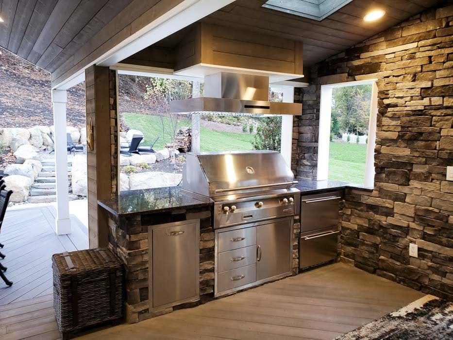 Elegant outdoor kitchen with stainless steel appliances and stone wall accents under a covered deck, overlooking a landscaped yard.