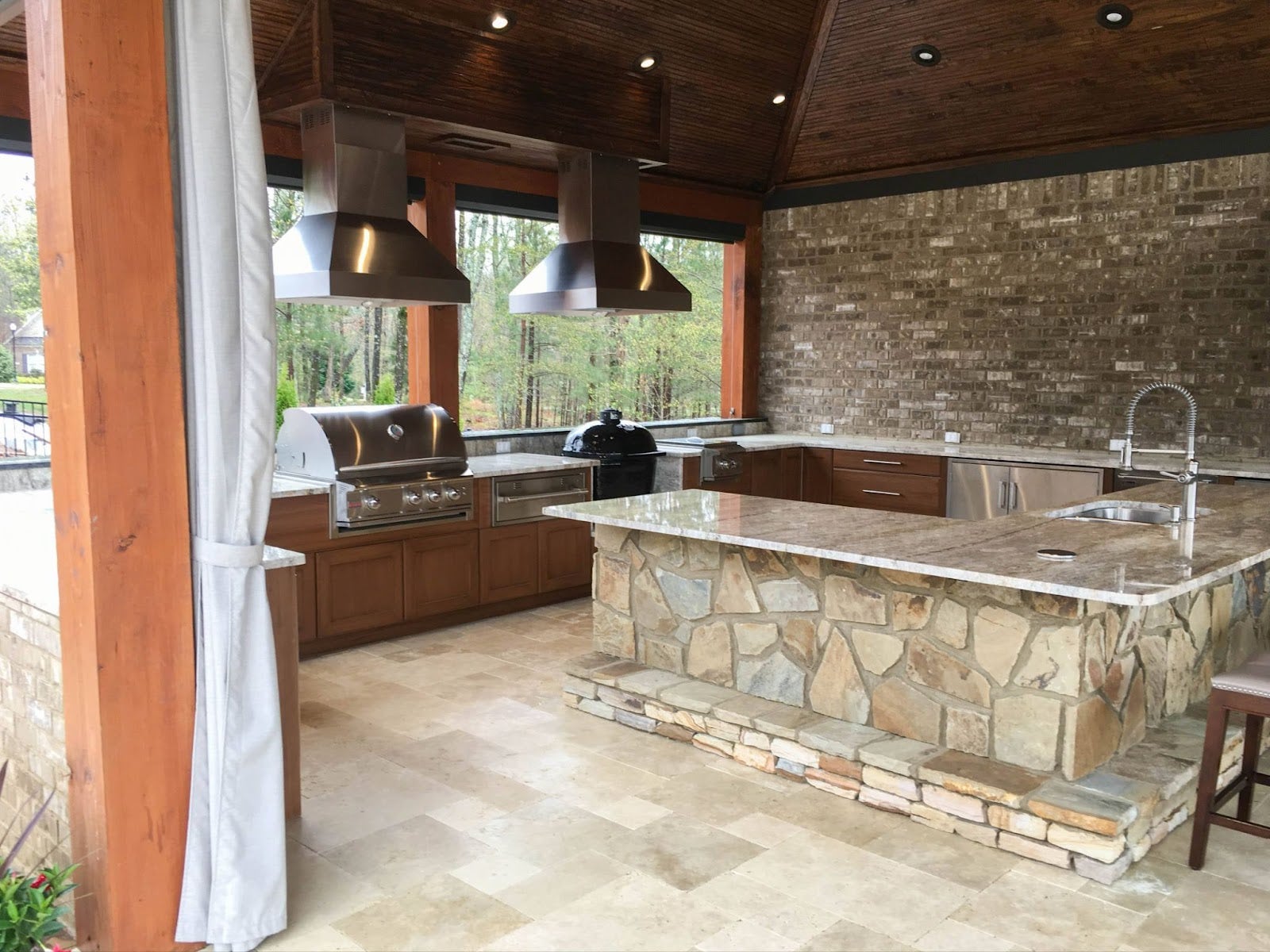 Expansive covered patio with a deluxe cooking area, including a grill and smoker under vent hoods, set against a brick wall with scenic views of nature.