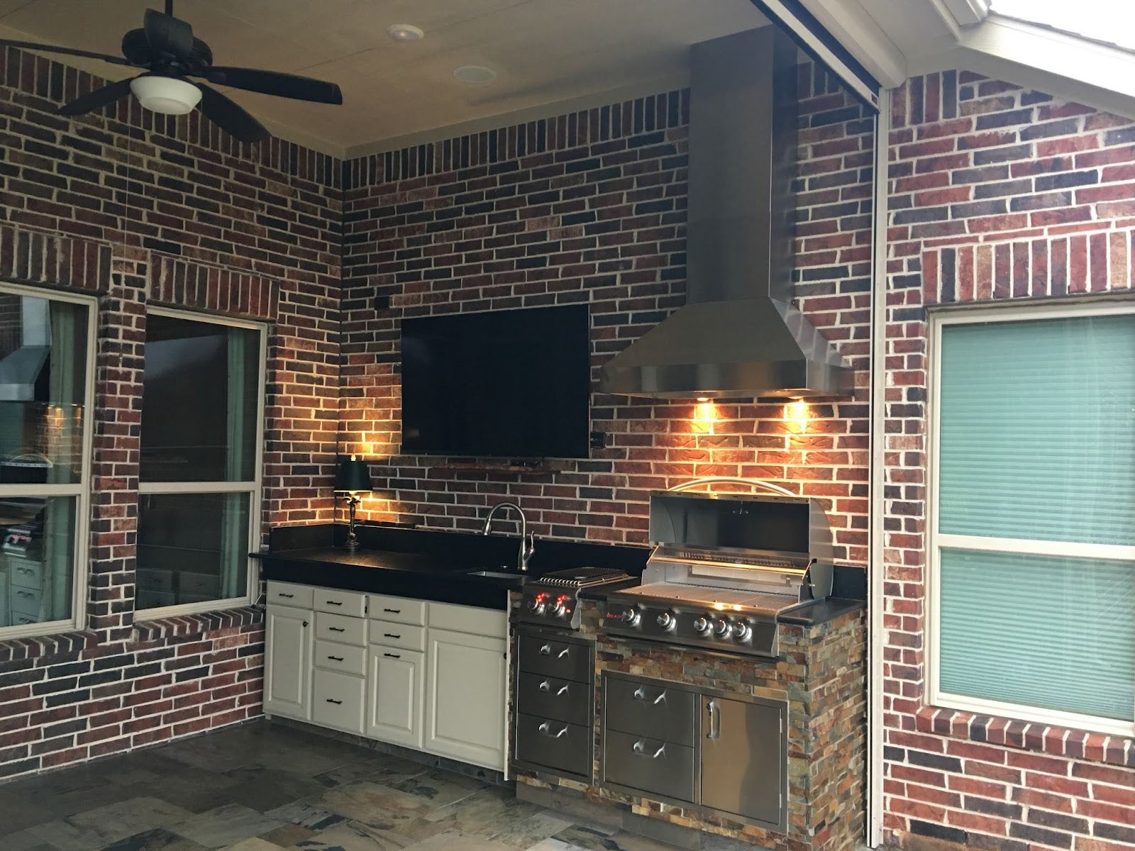 Enclosed patio with a classic brick wall, featuring a stainless steel Proline range hood over a grill and sink, complemented by ambient lighting - prolinerangehoods.com.