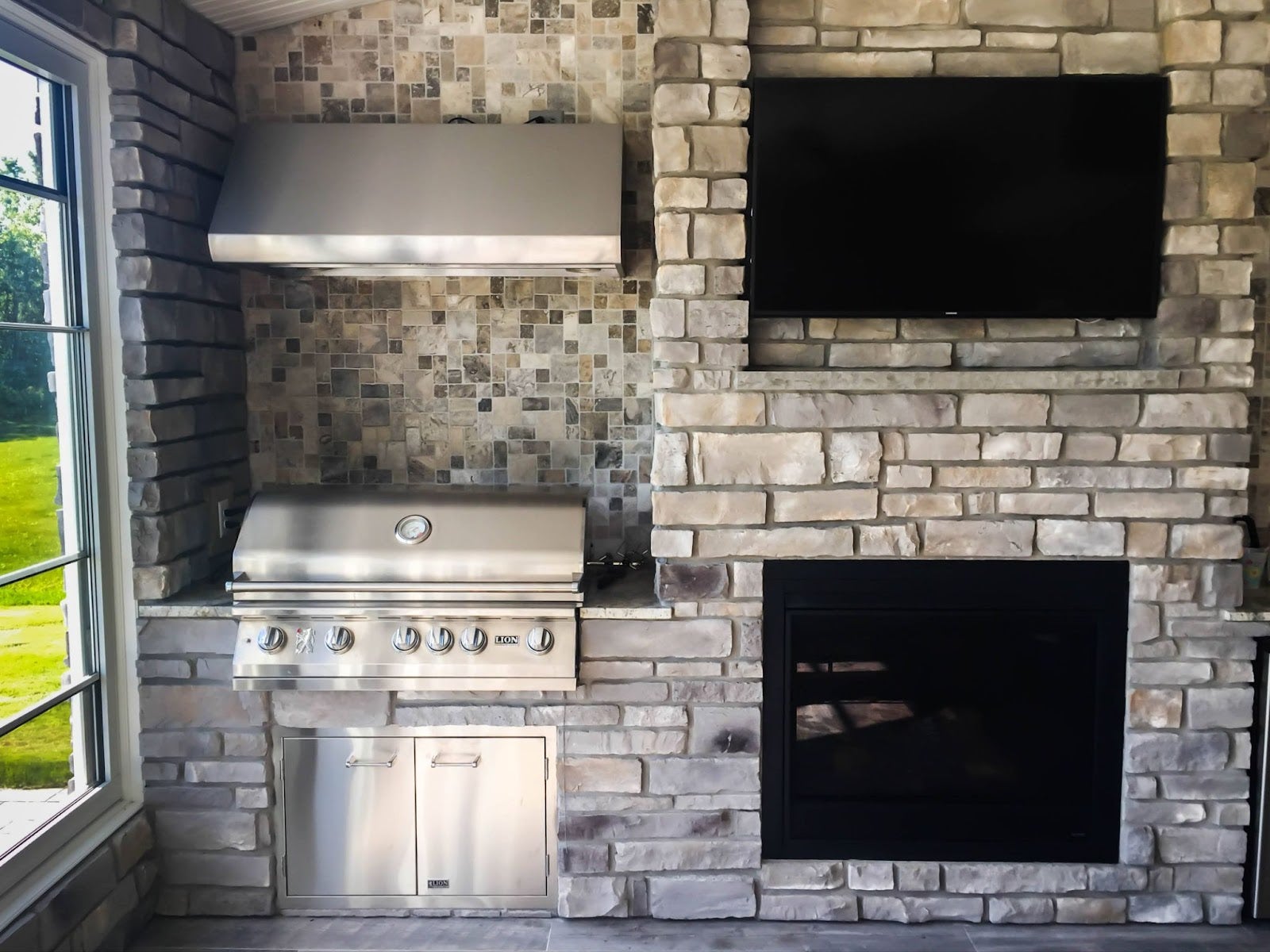 Luxurious outdoor kitchen setup with Proline range hood, matching grill, and stone fireplace under a flat-screen TV - prolinerangehoods.com