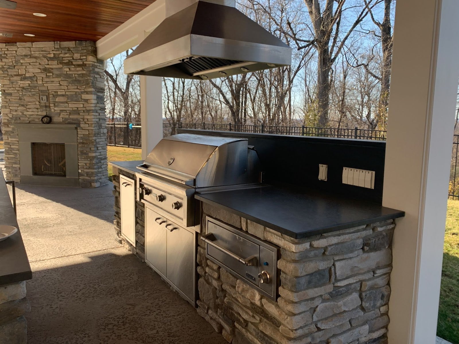 Elegant stone-clad outdoor cooking space with a professional stainless steel grill and matching ventilation system, adjacent to a fireplace.