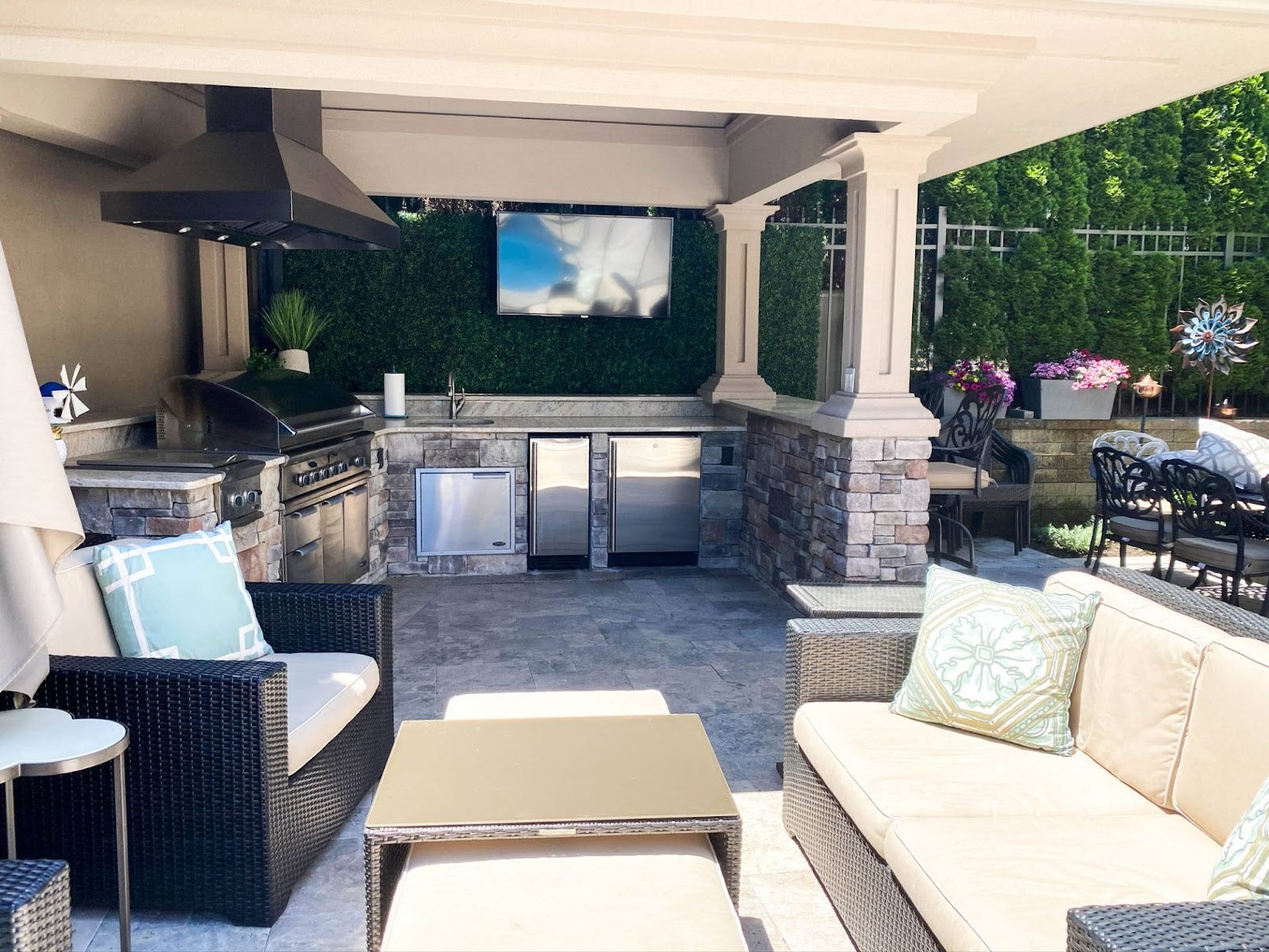 Inviting outdoor patio lounge area with comfortable seating and a fully equipped kitchen set against a green hedge.