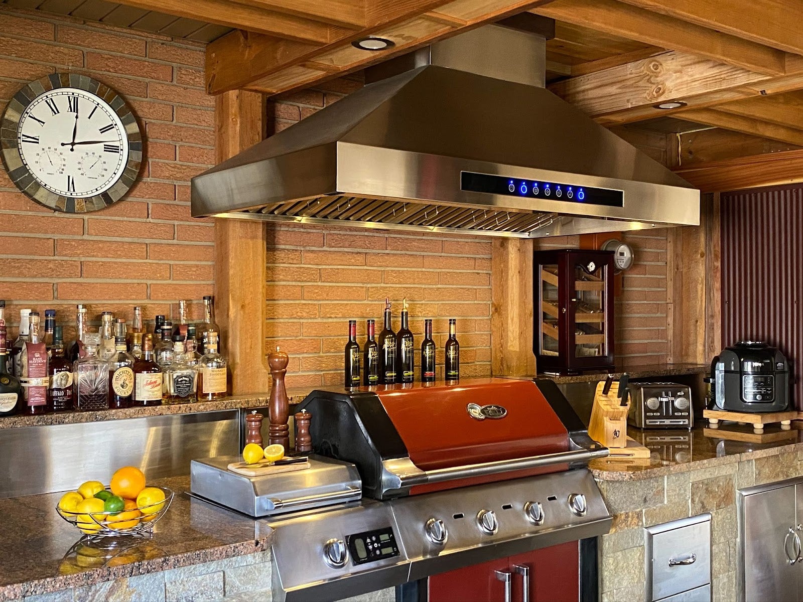 Home kitchen bar setup with a prominent stainless steel range hood above a red grill, complemented by a diverse alcohol selection.
