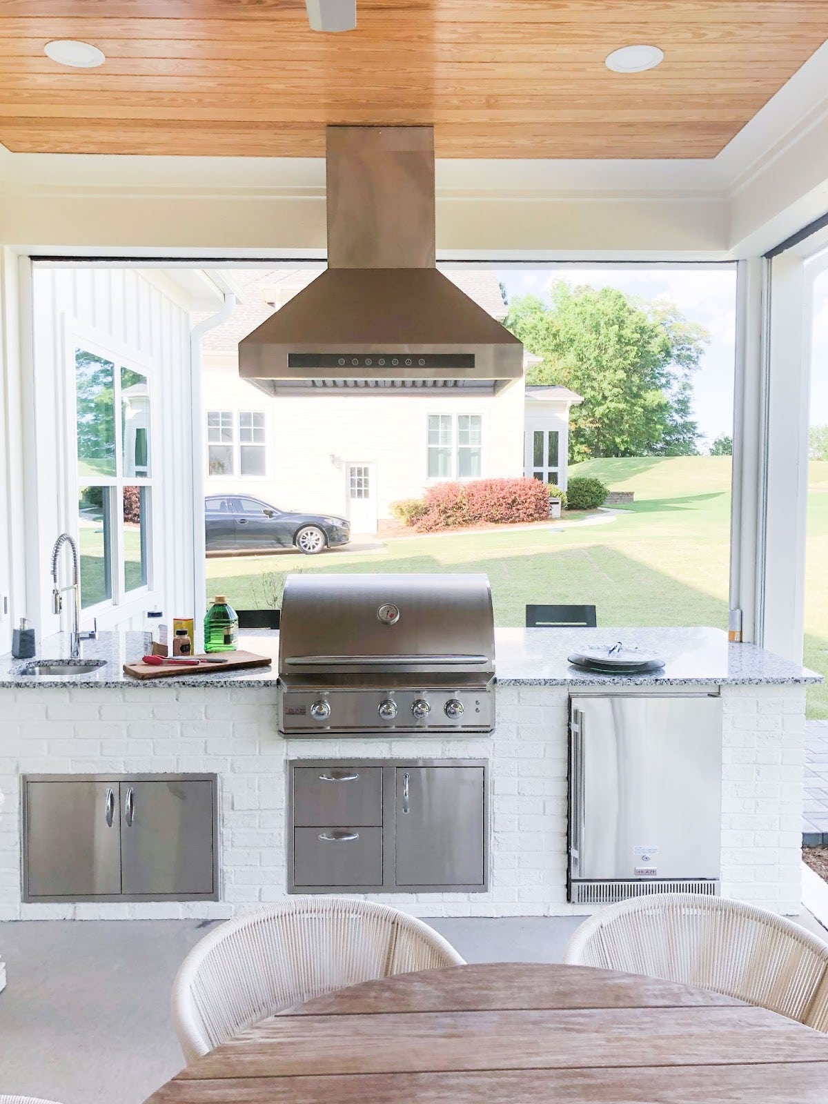 Sunlit patio kitchen with sleek appliances, including a grill and refrigerator, integrated into a clean white brick setting with a green lawn in the background.