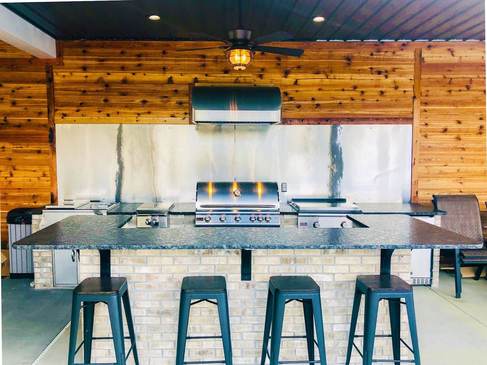 Outdoor entertainment space with a Proline range hood over a built-in grill, set against a backdrop of wood planks, brickwork, and a granite bar with stools - prolinerangehoods.com.