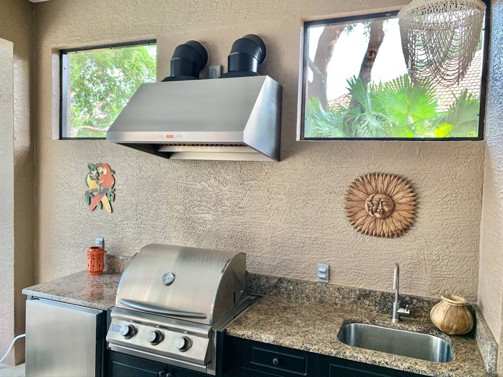 Cheerful outdoor grilling station with a Proline range hood, complete with a parrot wall ornament and sun face sculpture, creating a fun cooking space - prolinerangehoods.com.