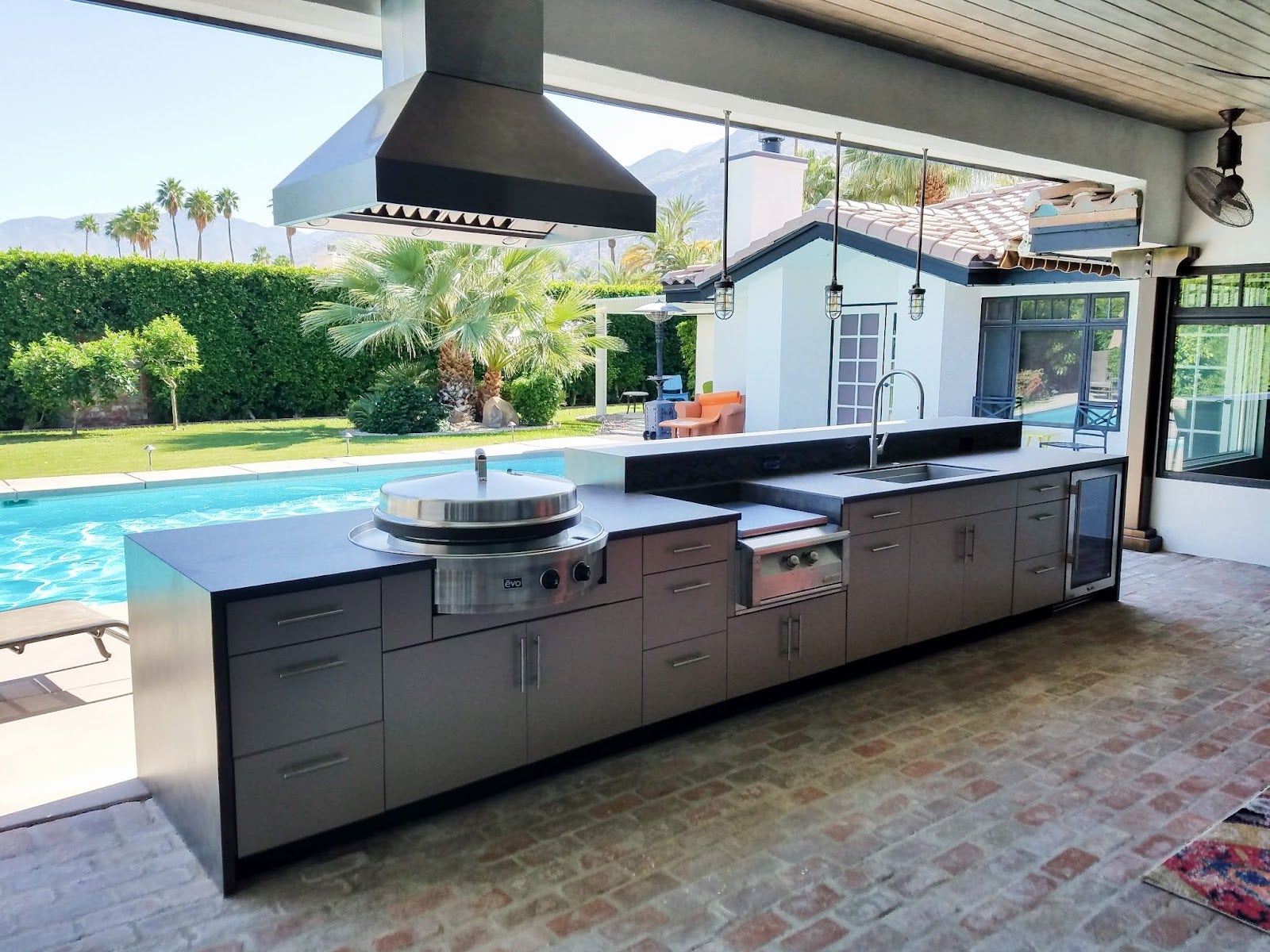 Resort-style outdoor kitchen setup with Proline ProVI range hood, complete with a pool and palm tree scenery in the background - prolinerangehoods.com