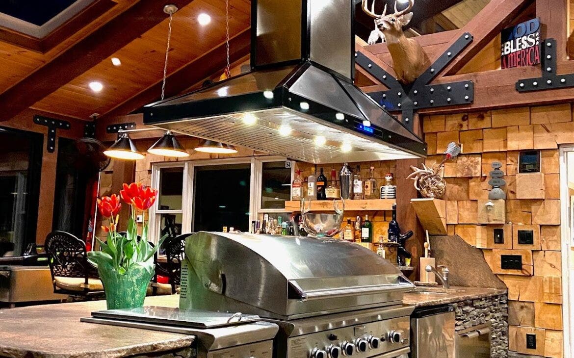 Warmly lit Proline range hood towering over an inviting outdoor kitchen with stone finishes - prolinerangehoods.com