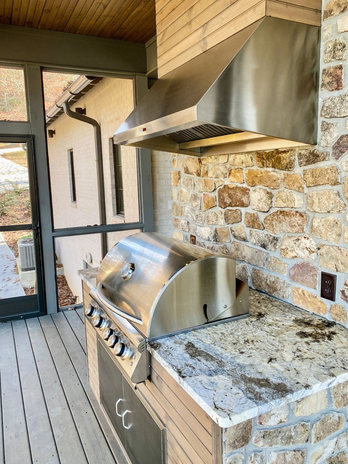 An elegant outdoor kitchen with a stainless steel range hood, built into a stone cooking area, surrounded by lush greenery -prolinerangehoods.com.