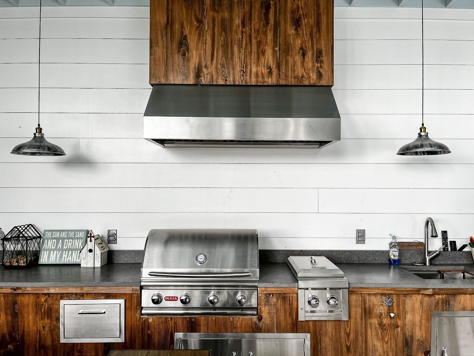 Outdoor kitchen blending rustic charm with modern functionality, featuring a Proline range hood and wooden cabinetry against a white shiplap wall - prolinerangehoods.com