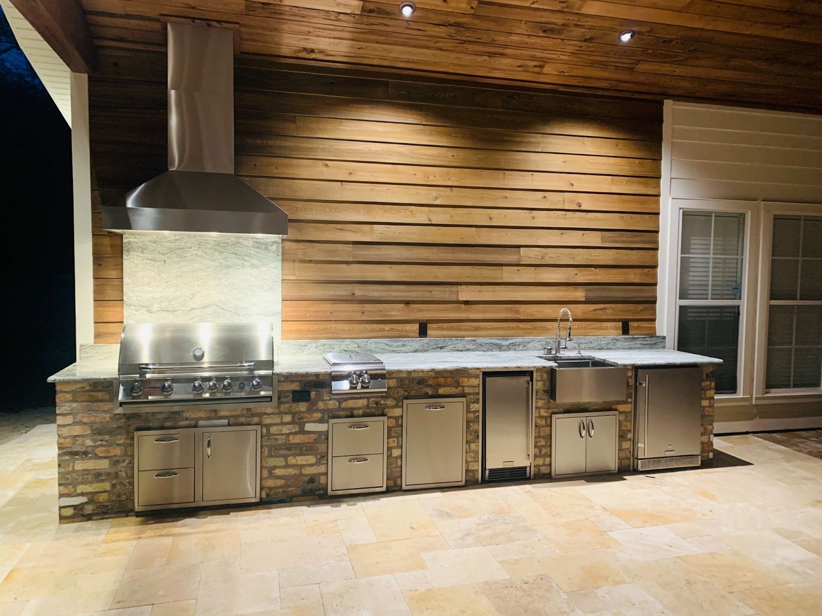 Rustic yet modern outdoor kitchen with stone counters, a Proline range hood, and a suite of stainless steel appliances under a warm wooden ceiling - prolinerangehoods.com.