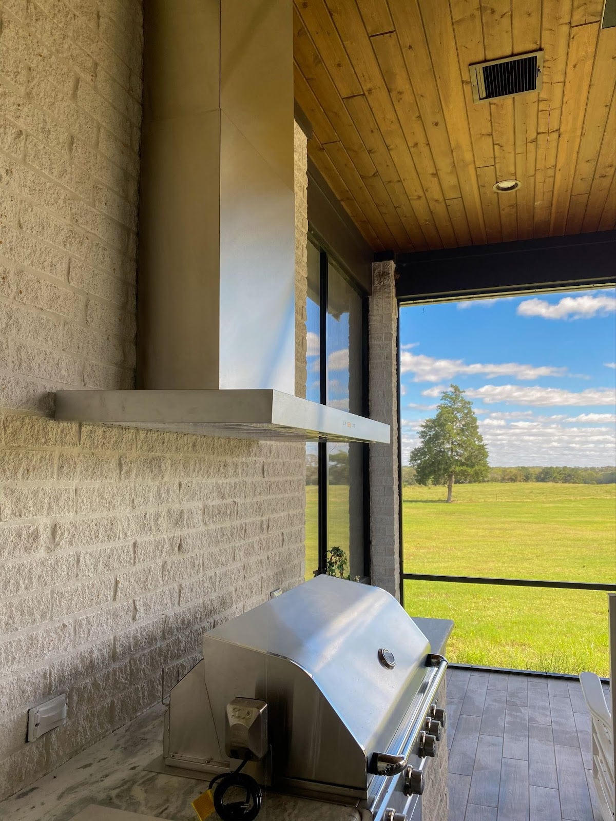 Inviting outdoor kitchen featuring a modern Proline range hood, stainless steel grill, and tranquil views of green fields under a wooden ceiling - prolinerangehoods.com