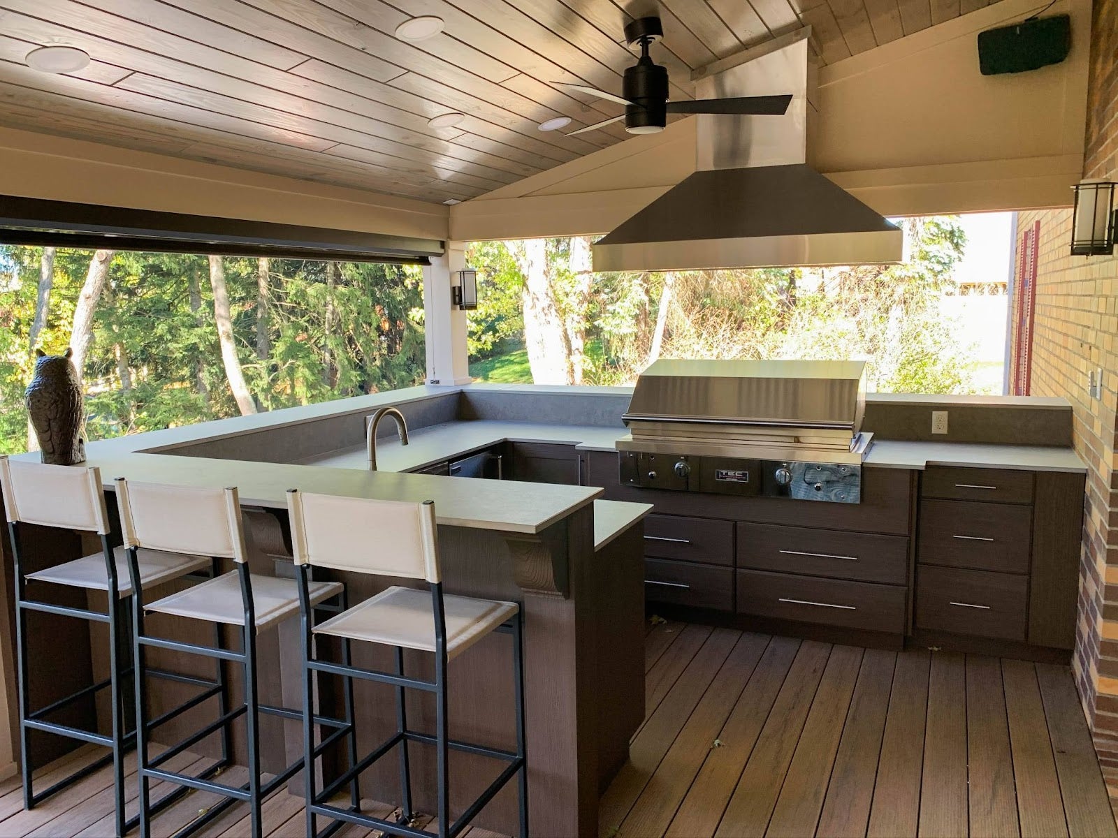 Outdoor kitchen with Proline range hood, bar stools, and wood decking nestled in nature - prolinerangehoods.com