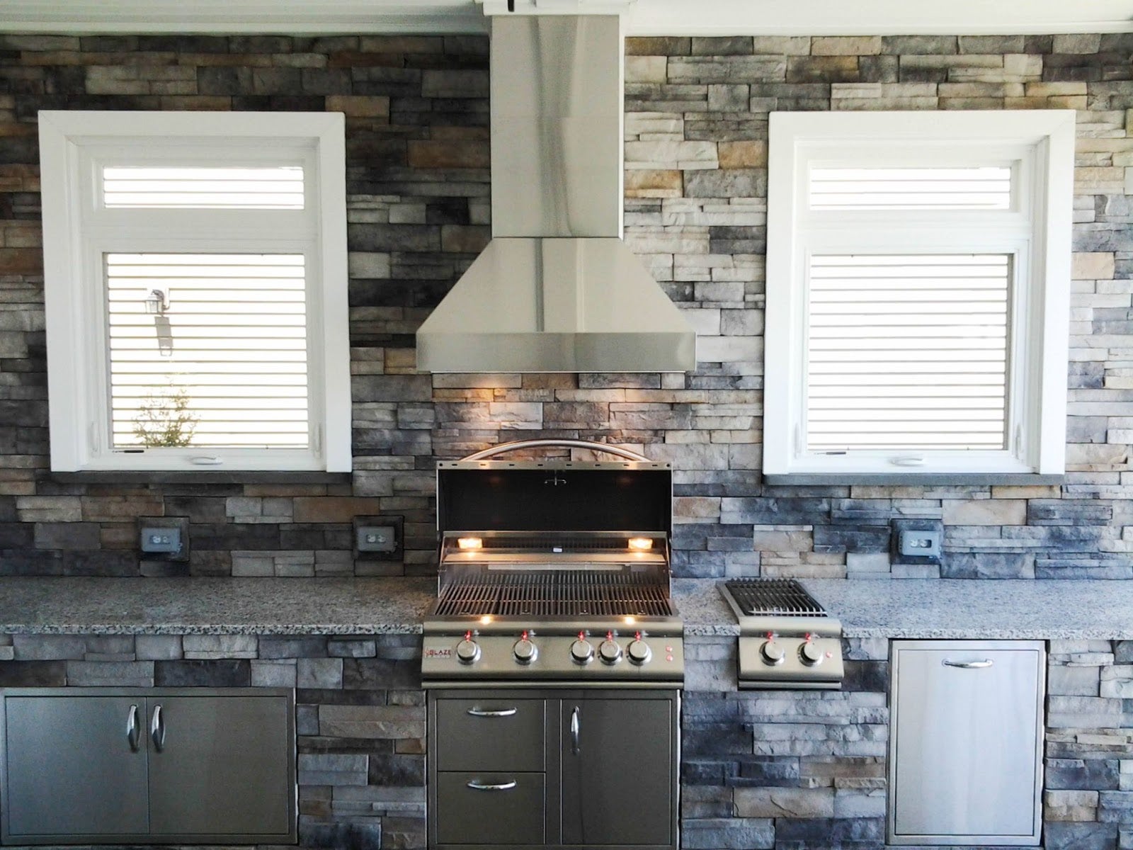 Sleek kitchen design with a stainless steel Proline range hood, matching grill, and granite countertops against a stone tile backsplash - prolinerangehoods.com.